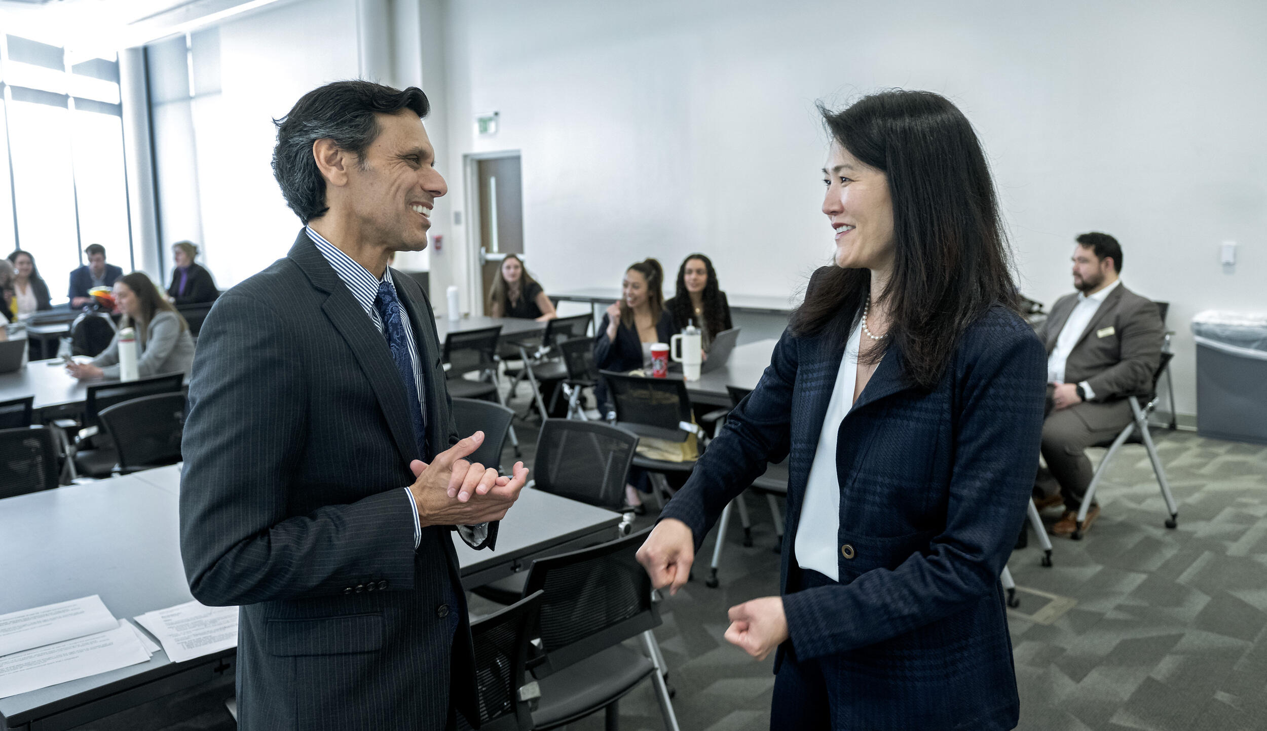 A photo of a mand and a woman speaking to each other in a room full of tables with people sitting at them. 