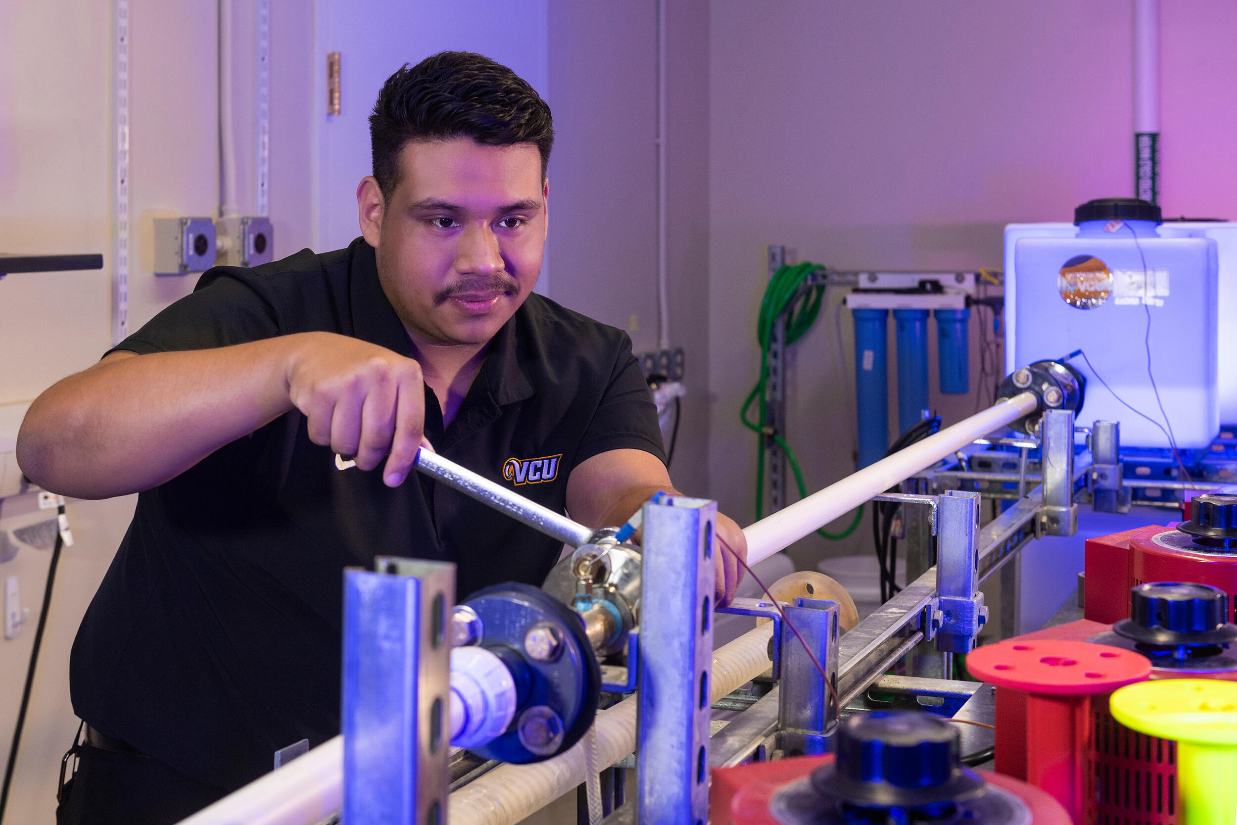 A photo of a man sitting at a table holding a wrench and working on an object made out of pipes.