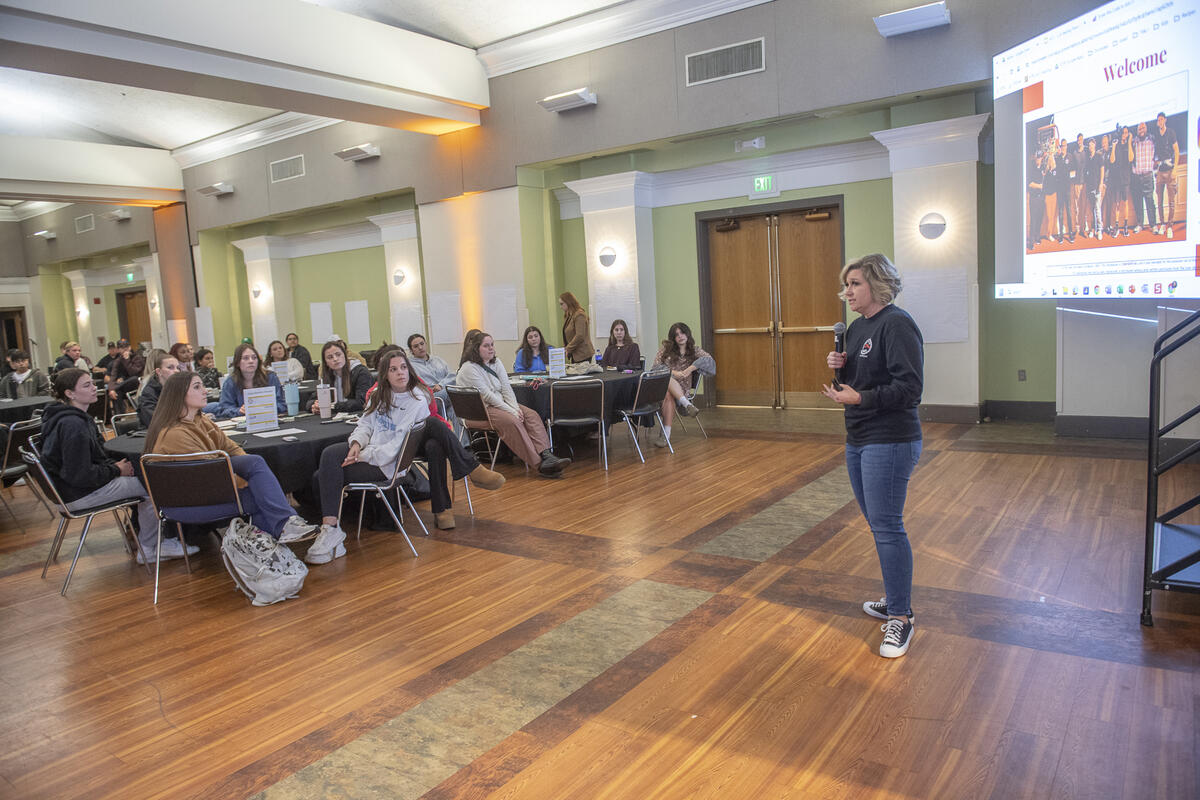 A photo of a woman speaking in a microphone to a room full of people sitting at tables. 