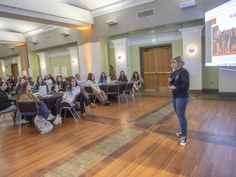 A photo of a woman speaking in a microphone to a room full of people sitting at tables. 