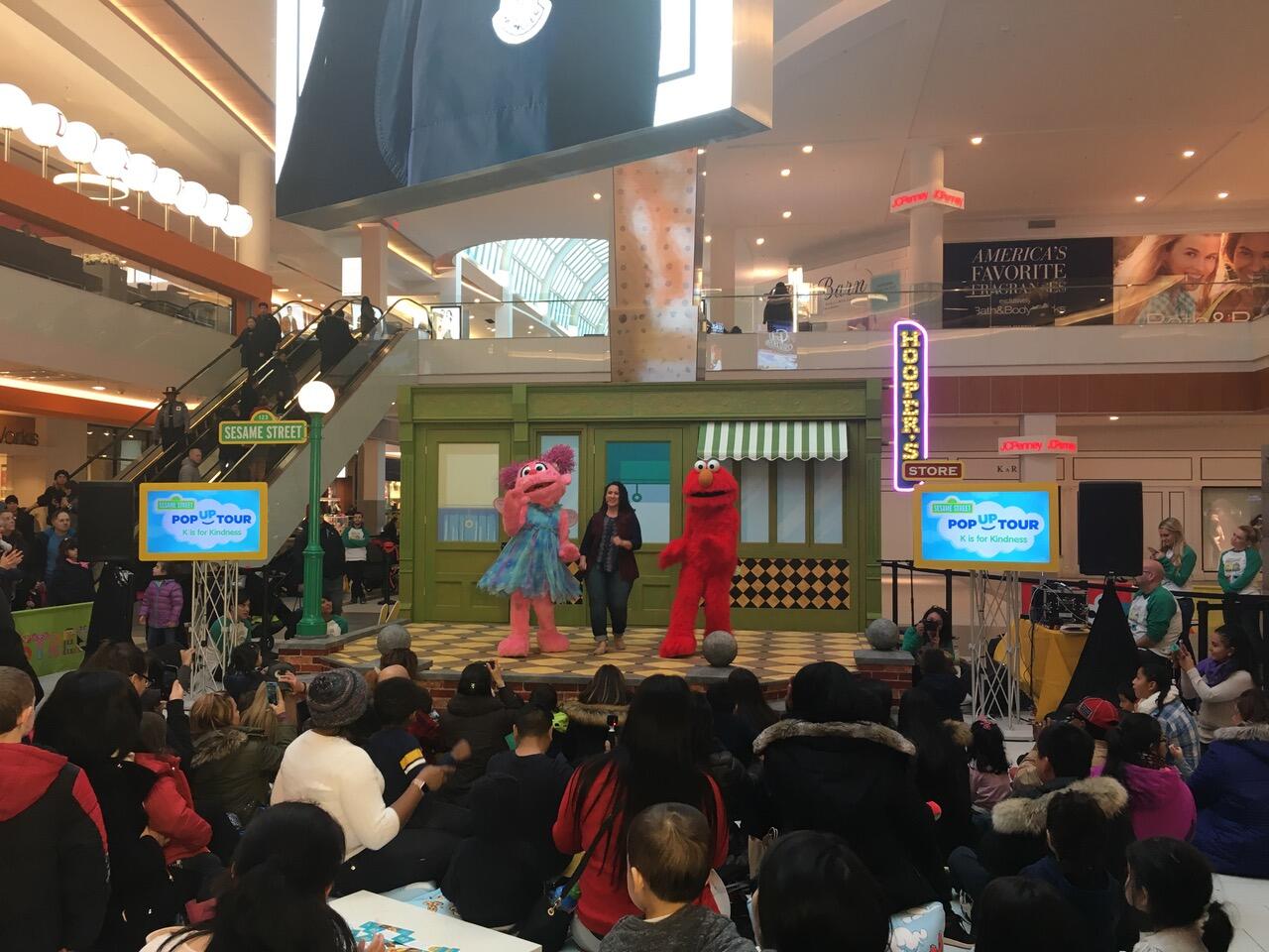 A photo of a crowd of people at a mall surrounding a stage. On the stage are two people in Sesame Street mascot costumes and one woman. Behind them is a set piece that looks like the front of a store. 
