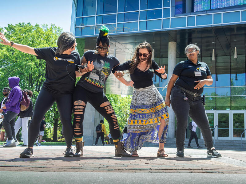 A group of VCU community members dance together in front of the new glass facade of a building on a sunny day on a brick sidewalk.
