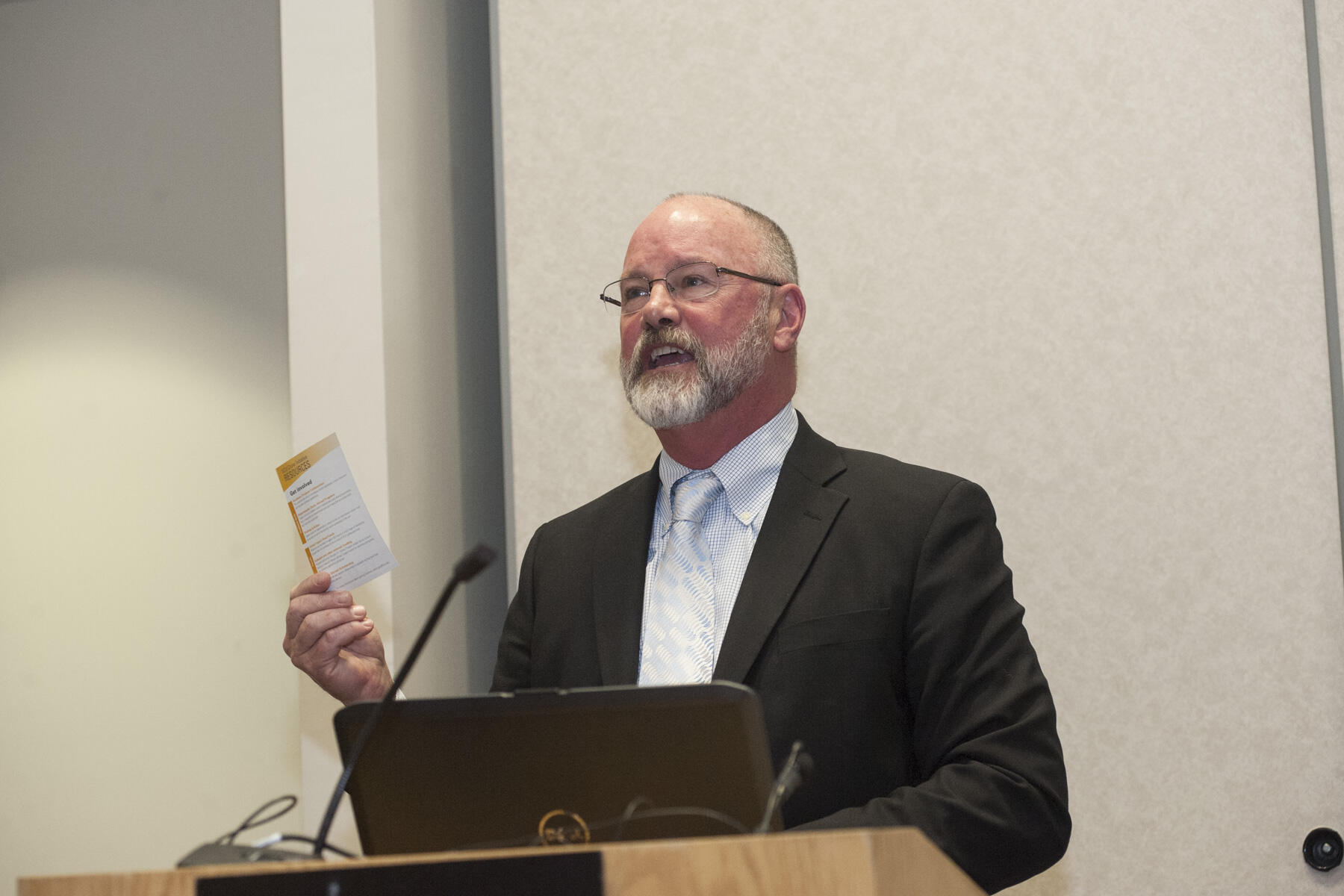 A man wearing a suit standing at a podium speaking while holding a small piece of paper in his left hand. 