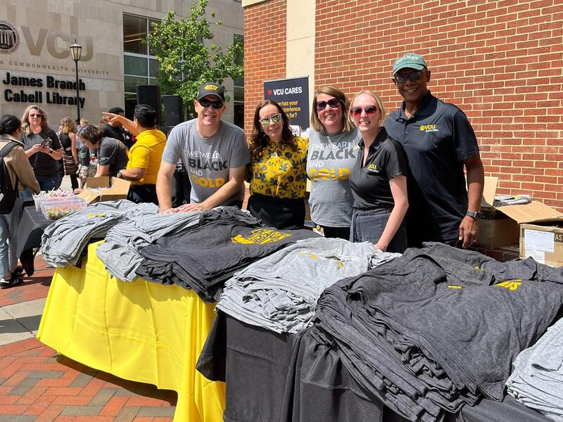 A photo of five people standing behind two tables with yellow and black table cloths. The tables have stacks of T-shirts on them. 