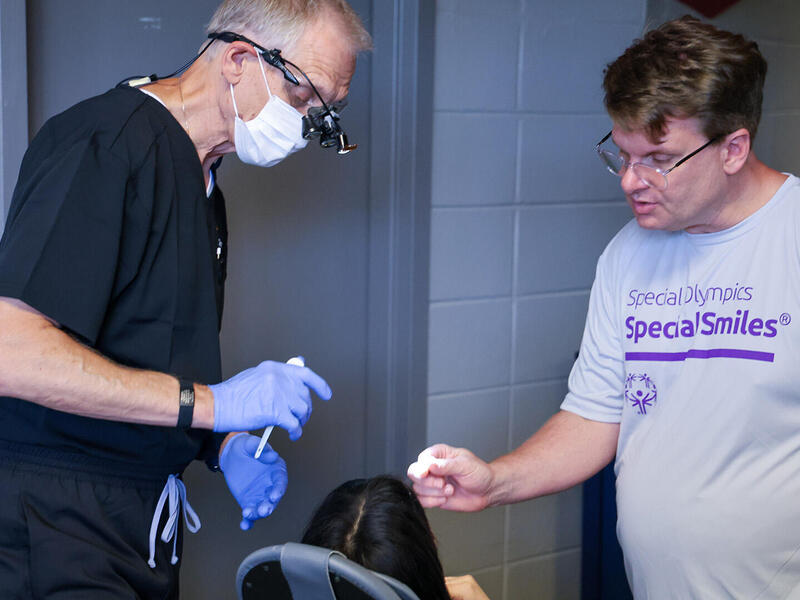 A photo of two men standing over a person in a dental chair. The man on the left is wearing dental scrubs, blue latex gloves, a face mask, and magnifying glasses. The man on the right is wearing glasses and a t-shirt that says \"Special Olympics Special Smiles\" in purple text. 