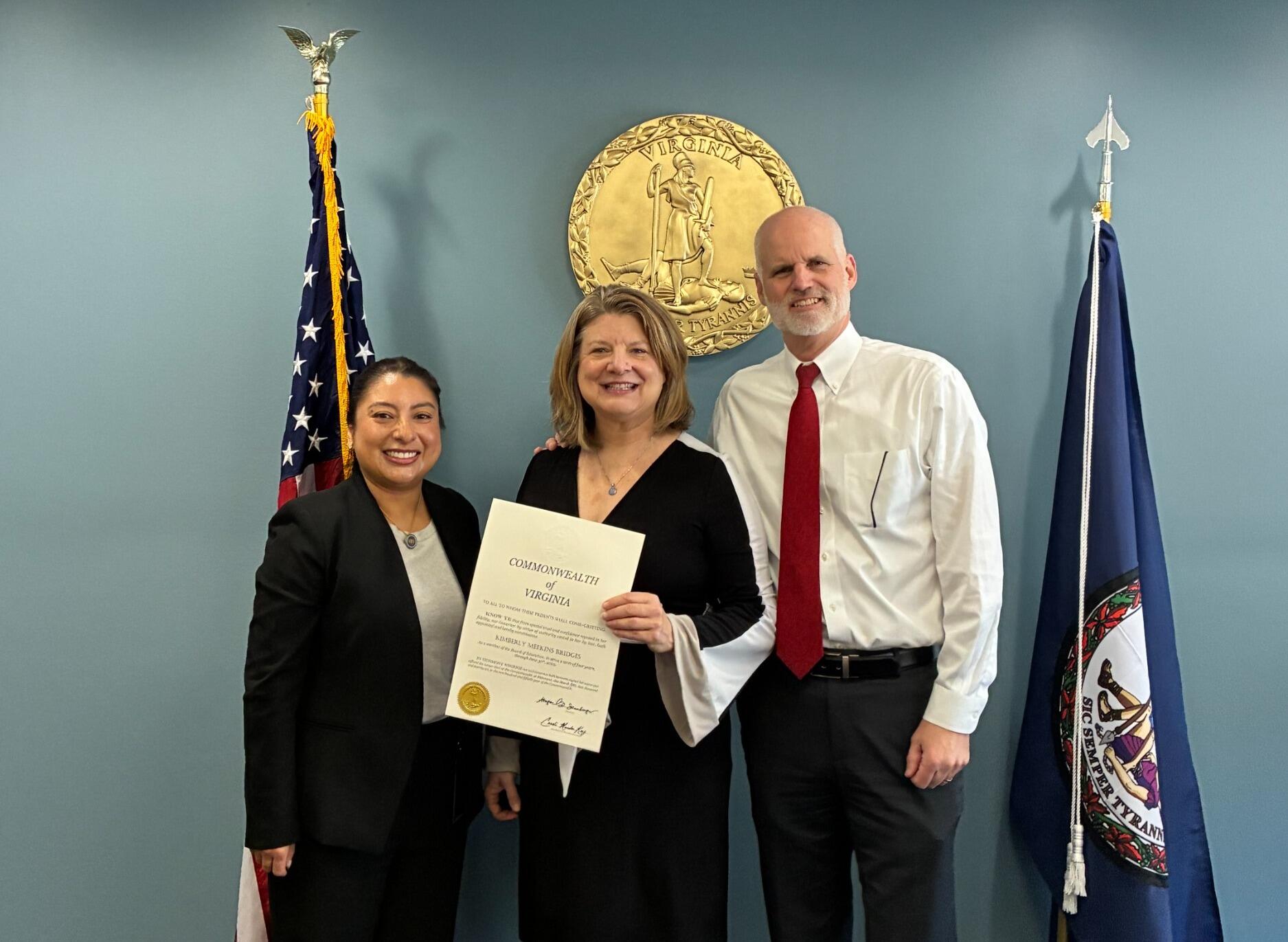 A photo fo three people standing. The person in the middle is holding a certificate. 