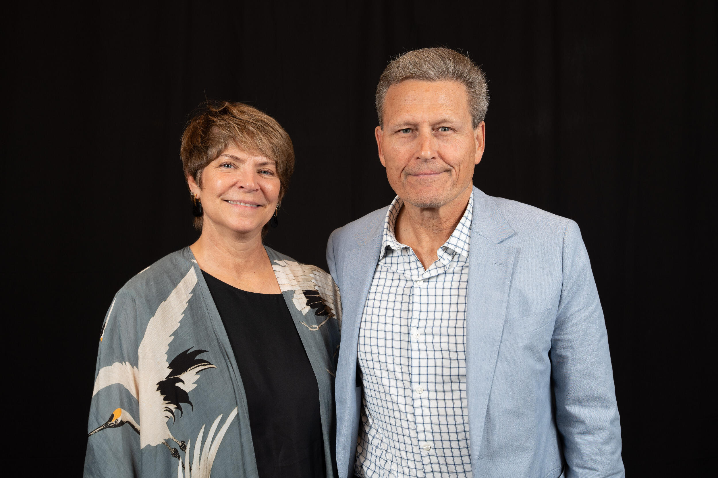 A woman in a black shirt and sweater and a man in a white shirt and suit jacket stand shoulder to shoulder.