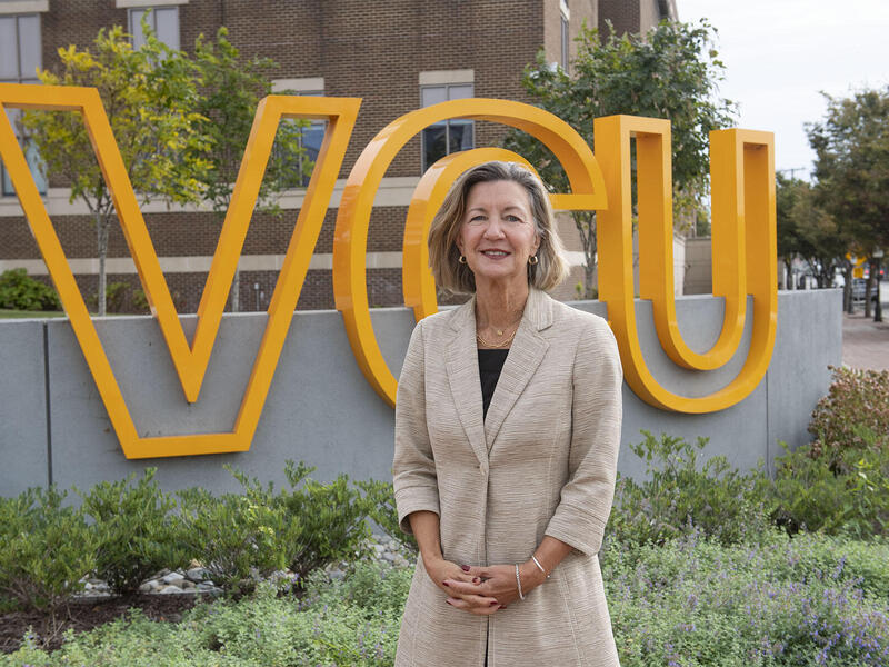 A photo of a woman from the hips up standing in front of a sign with giant yellow letters that spell out \"VCU\"