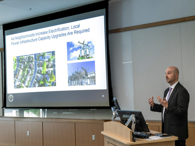 A man stands at lectern and speaks with powerpoint behind him.