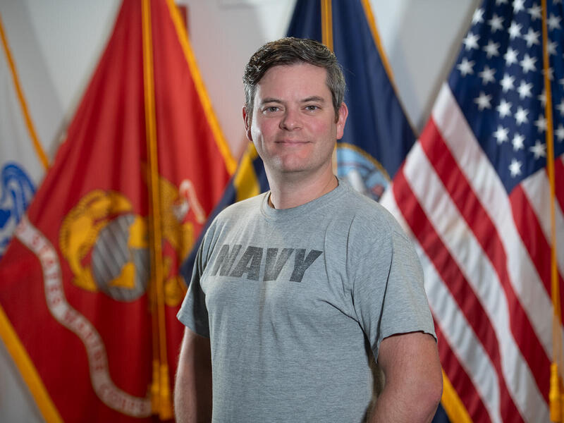 Man in a Navy T-shirt stands in front of two flags.
