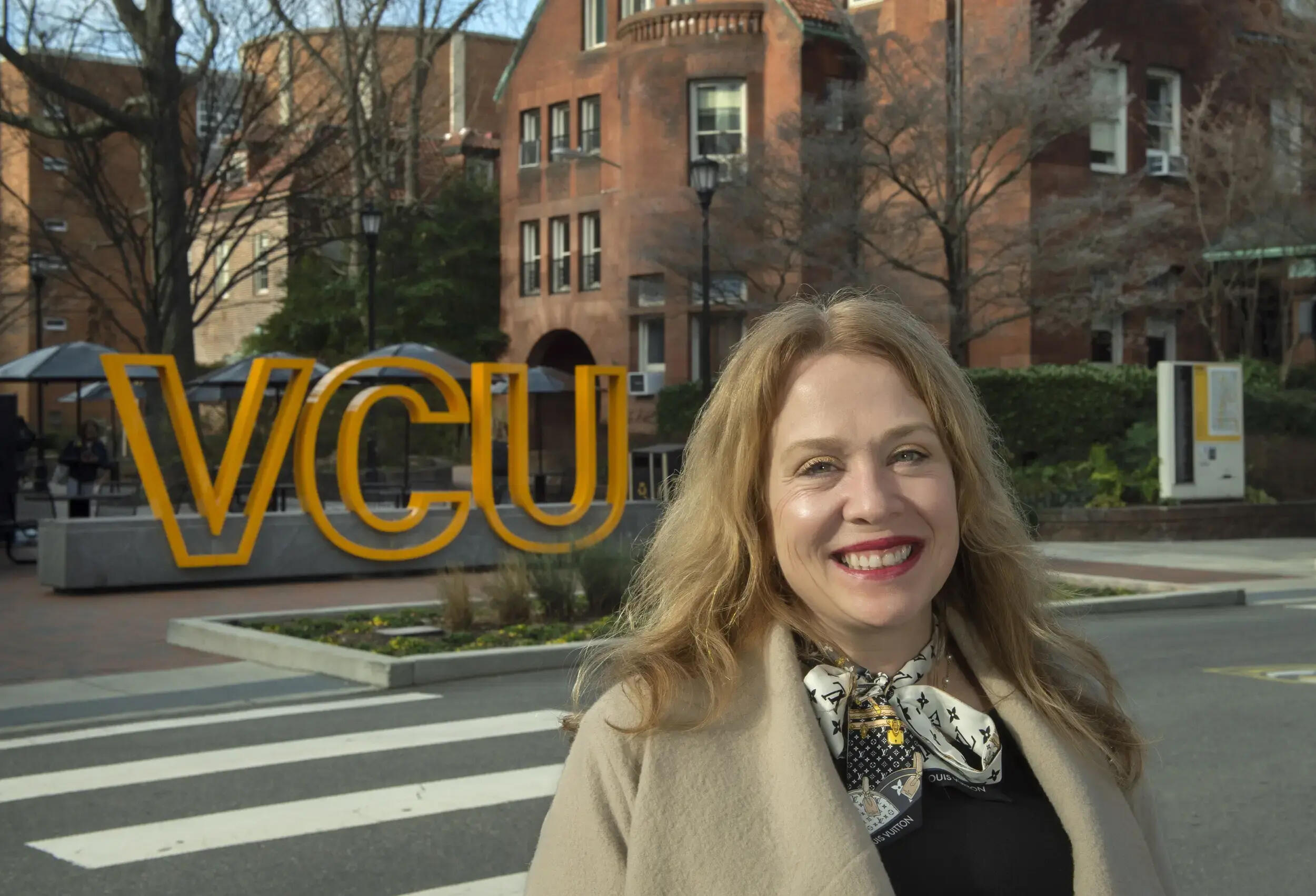 A photo of a woman from the chest up. Behind her is a sign made up of large yellow letters that spell out \"VCU.\" 