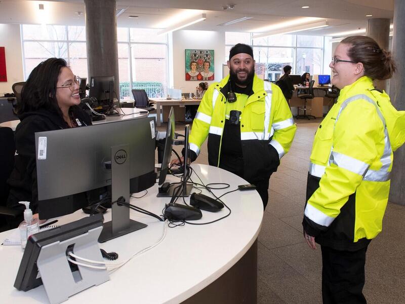 A woman at a desk with two computers smiling at two people wearing yellow jackets with reflective material. 
