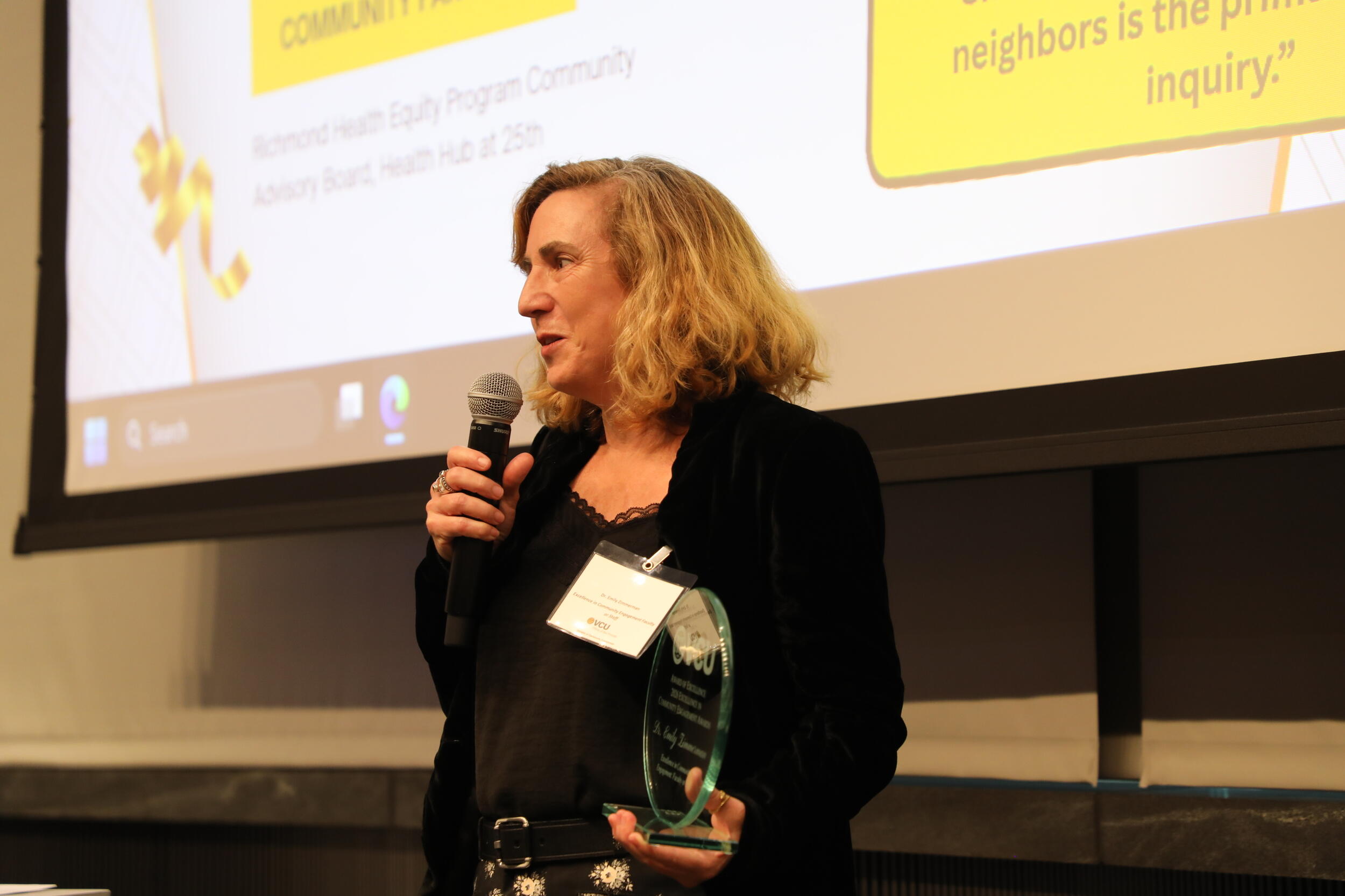 A photo of a woman from the waist up. She is speaking into a michrophone which she is holding with her left hand. In her right hand is a glass award trophy. 