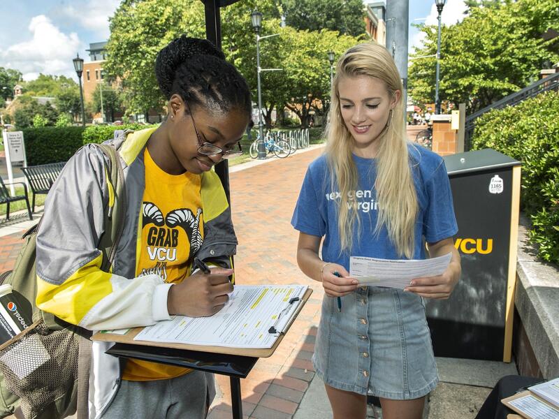A woman filling out a form on a clipboard. To her right is another women standing while holding some papers. They are both looking at the clipboard. 