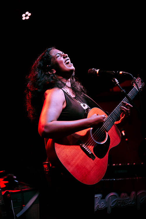 A woman playing a guitar, leans back and sings into a microphone.