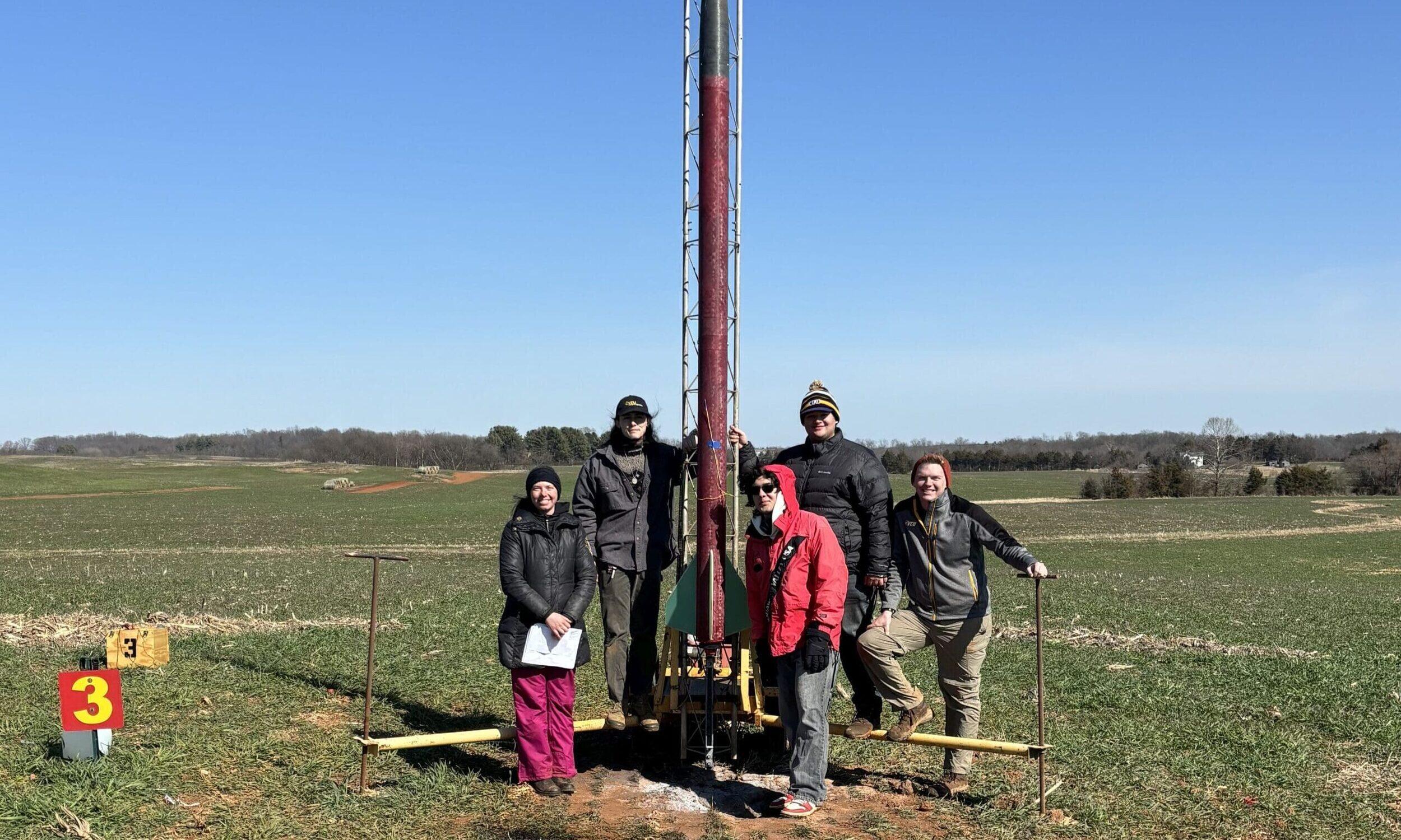A photo of five people standing in a field around a large pole 