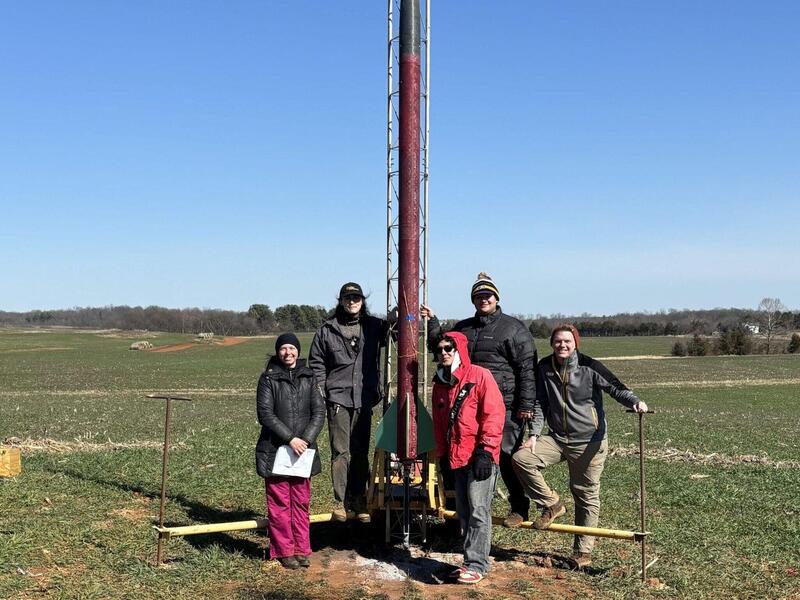A photo of five people standing in a field around a large pole 