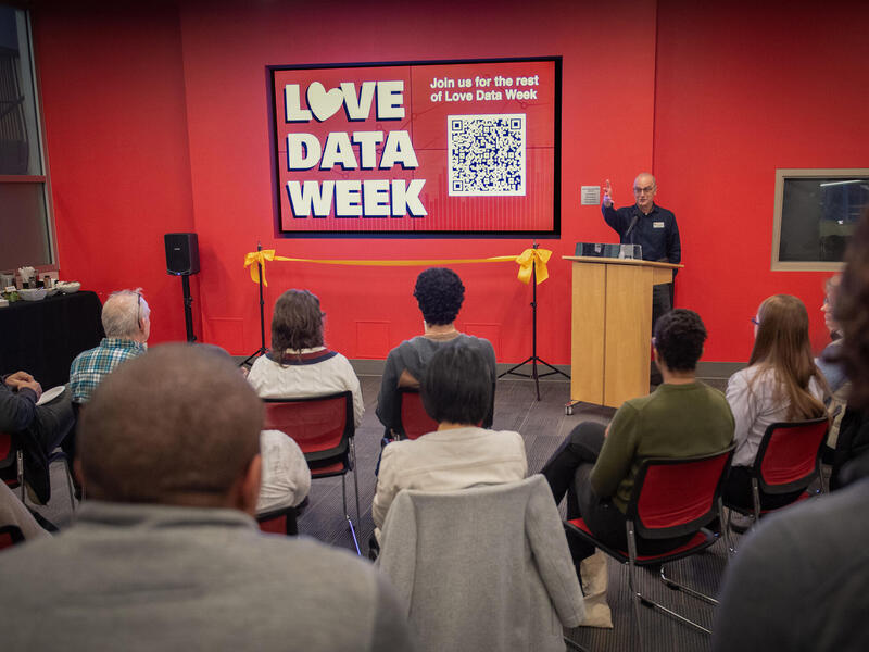 A photo of a room of people looking at a wall with a sign that says \"LOVE DATA WEEK\" in white letters. To the right of the sign is a man standing behind a wood podium and speaking. 