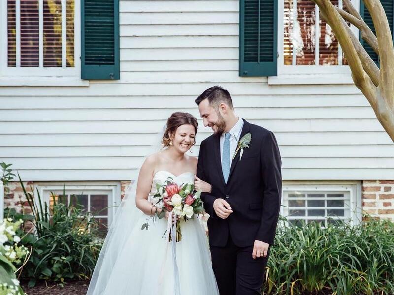 A photo of a woman wearing a wedding dress standing next to a man in a suit and tie. Her right hand is around the man's left arm, and her left hand is holding a wedding bouquet. They are standing in front of a white house with green shutters. 