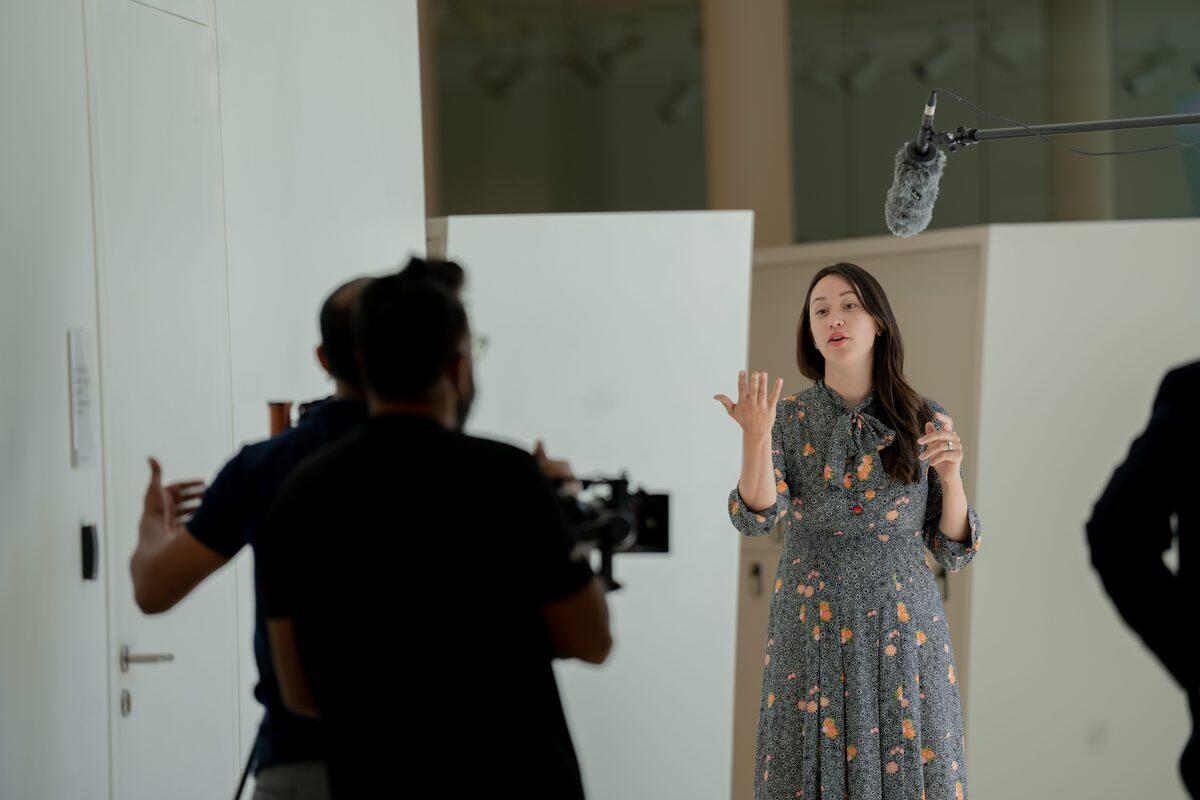 A man behind a camera filming a woman speaking. 