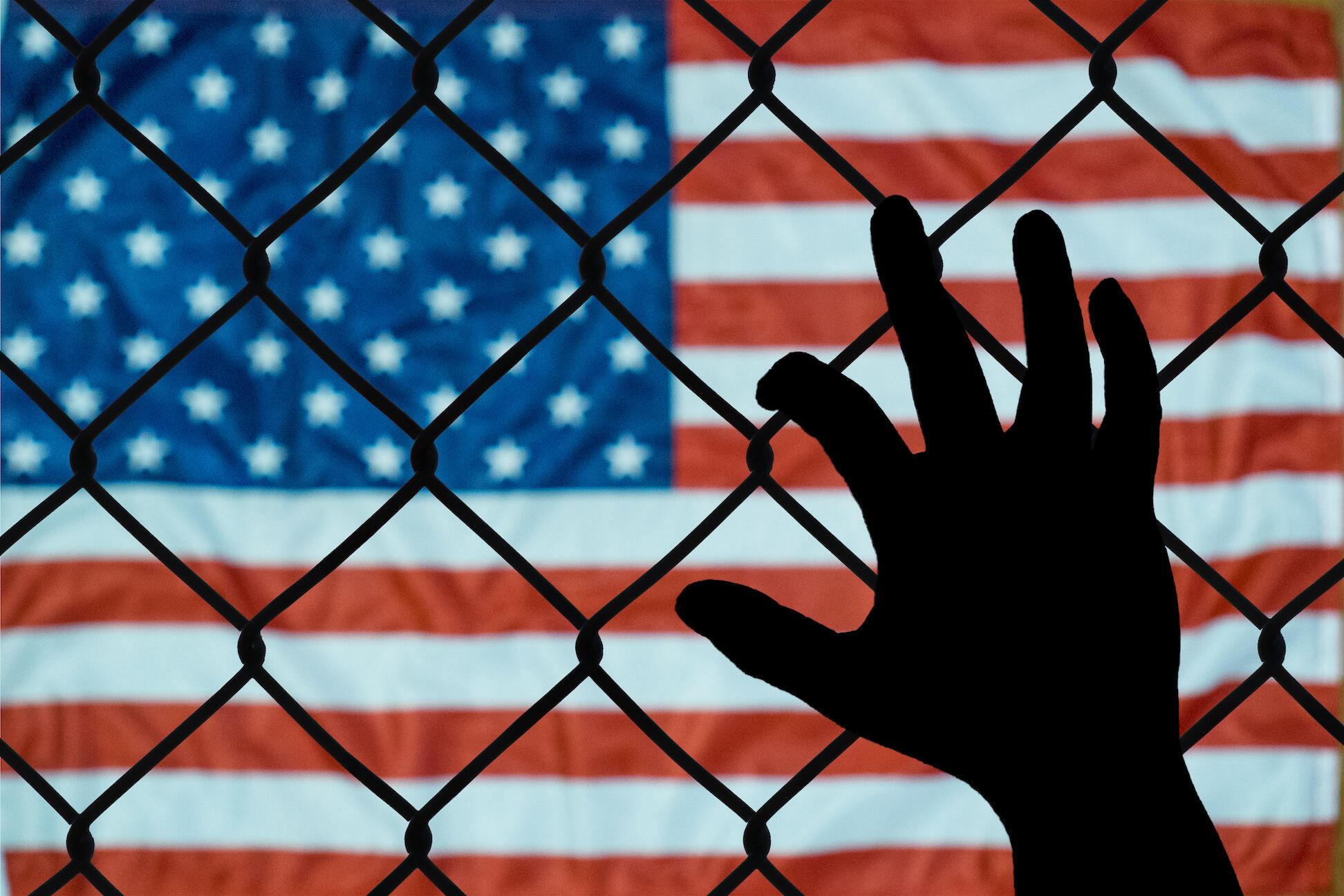 A hand grabs onto a wire fence. The flag of the United States of America is in the background.