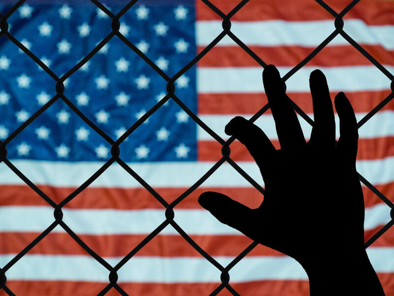 A hand grabs onto a wire fence. The flag of the United States of America is in the background.