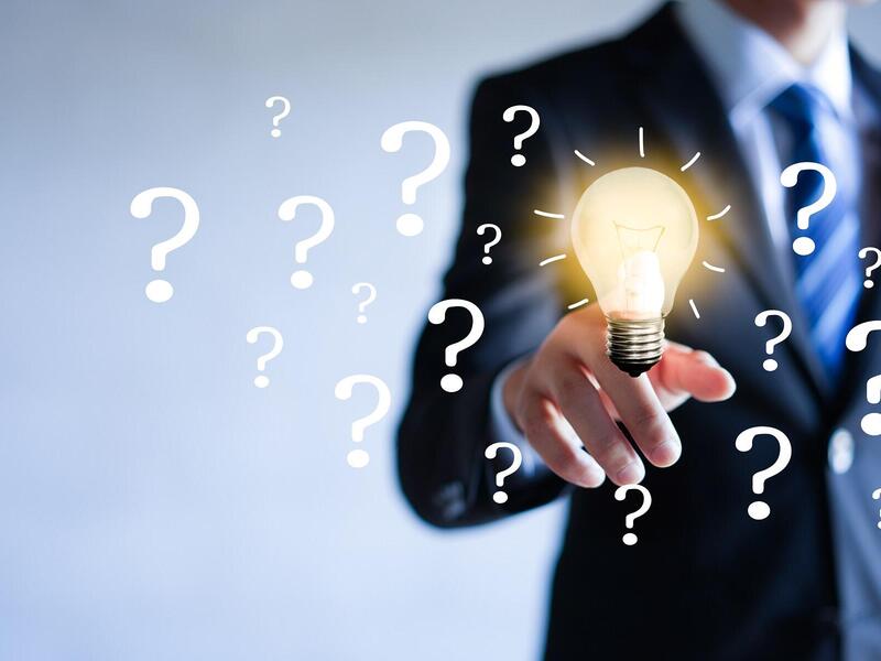 A person in business attire points at a lit light bulb surrounded by question marks.