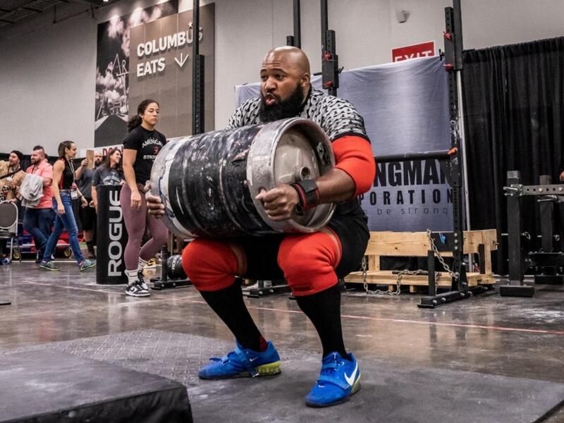 Three photo side by side that show a man going thorugh the process of lifting a barrel up in the air above his head. In the first photo the man has bent over to pick up the barrel, in the second photo he is squating holding the barrel, and in the thrid photo he is standing with the barrel above his head. 