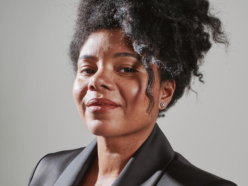 A photo of a woman from the shoulders up. She is wearing a coal gray sports coat and an off white blouse. 