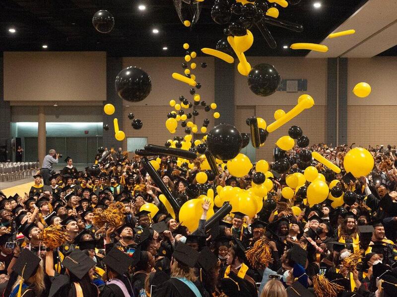 A crowd of graduates wearing graduation caps and gowns with red and yellow balloons falling down on them. 
