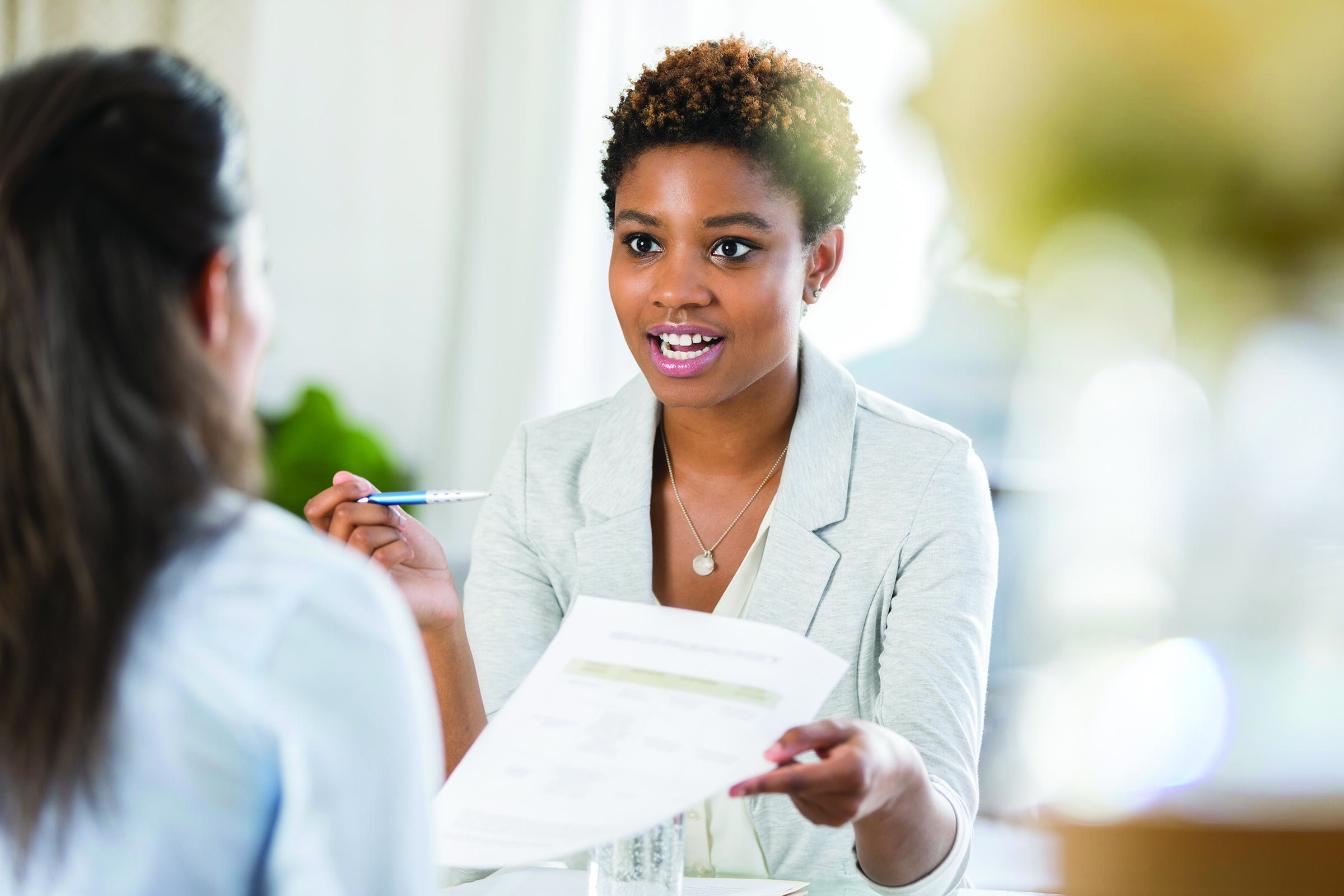 A person holding a pen and sheet of paper speaking to another person.