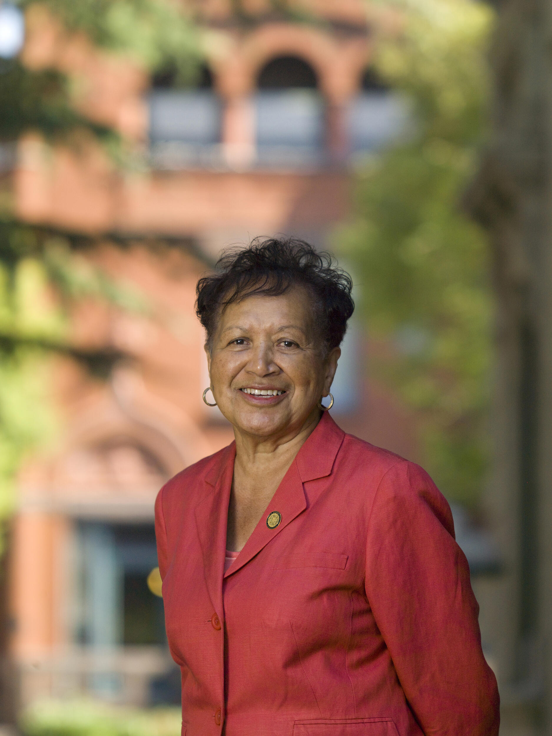 A photo of a woman from the chest up. She is wearing a red sports coat and is standing outside in front of a brick building. 