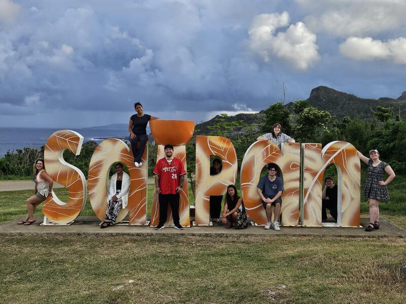 A group of people pose with a large sign spelling out SAIPAN in a field.