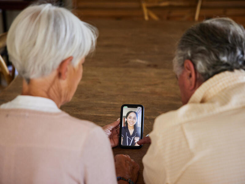 A photo of two elderly people holding a smart phone that has a video call with a medical care provider. 