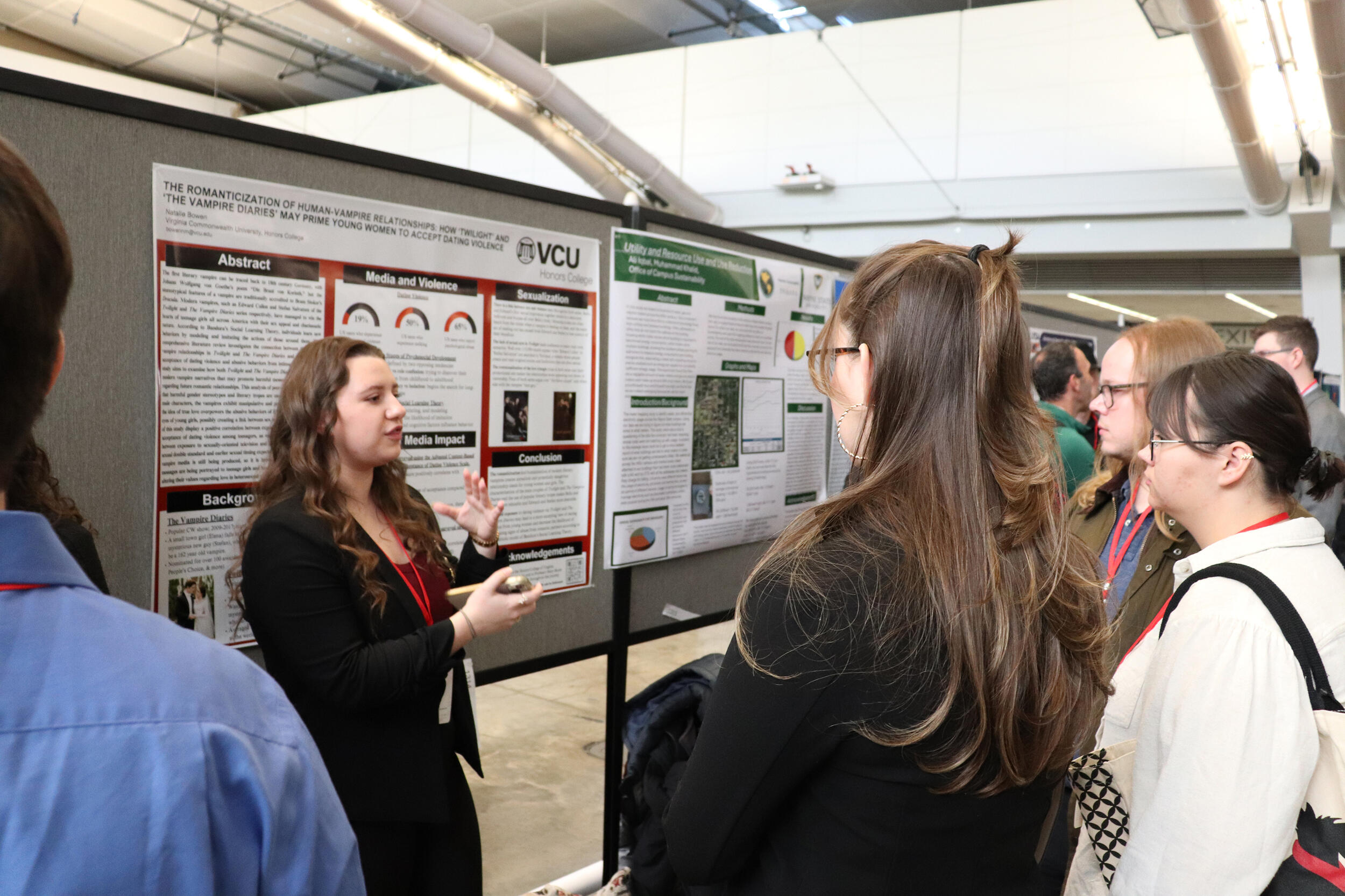 A photo of a woman standing in front of a poster and speaking. There is a crowd of people standing in a semi-circle listening to her speak. 