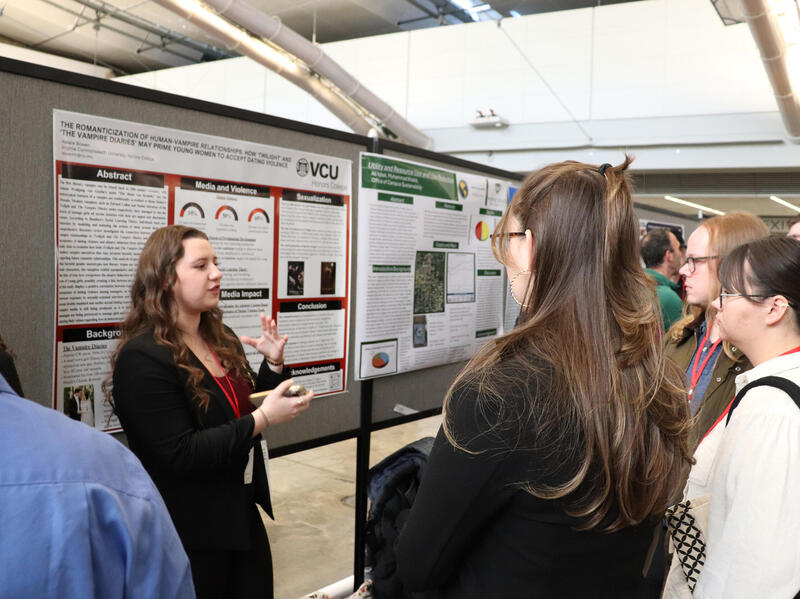 A photo of a woman standing in front of a poster and speaking. There is a crowd of people standing in a semi-circle listening to her speak. 
