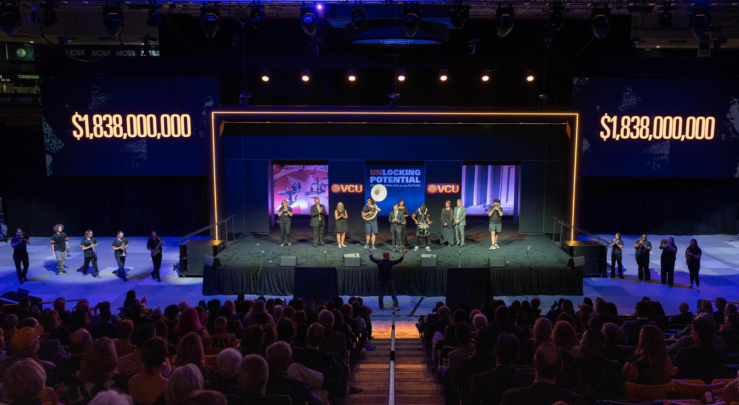 A photo of a stage sitting on a court. There are five people standing to the left of the stage, four standing to the right of the stage, and ten people standing on the stage. On either side of the stage are light up signs that read \"$1,838,000,000.\"