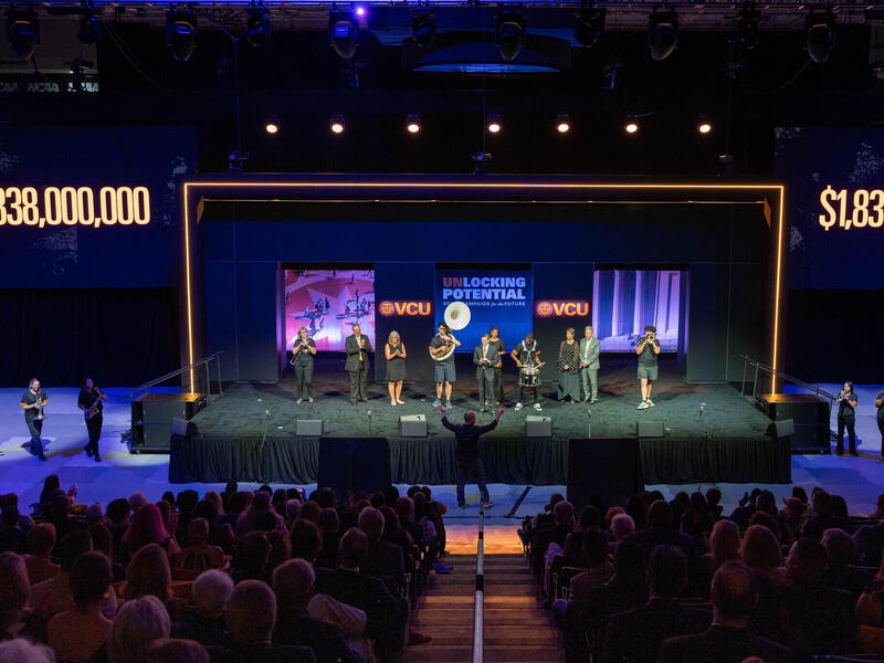 A photo of a stage sitting on a court. There are five people standing to the left of the stage, four standing to the right of the stage, and ten people standing on the stage. On either side of the stage are light up signs that read \"$1,838,000,000.\"