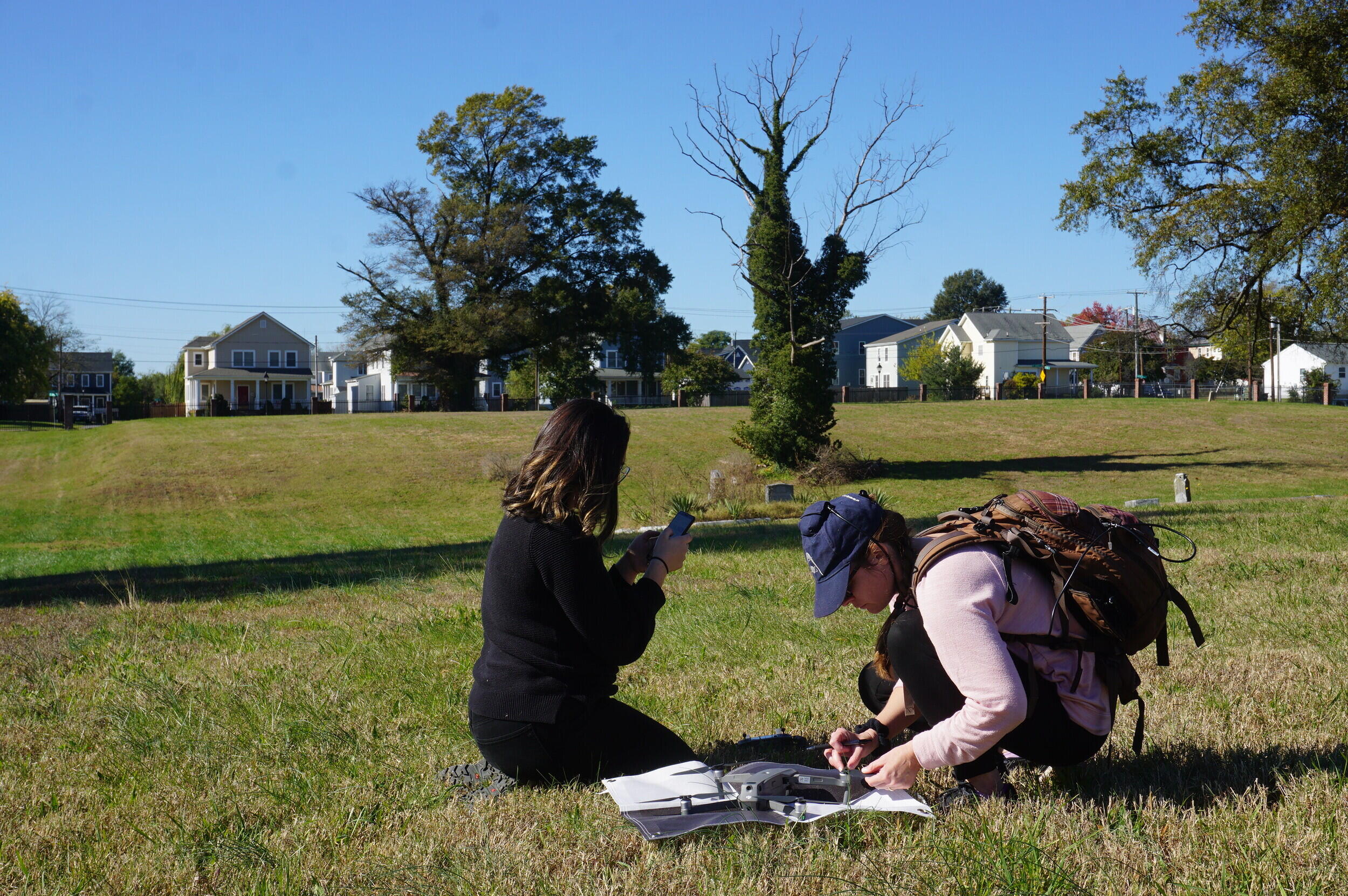 Two women kneeling in a grassy area with papers and books in front of them. 