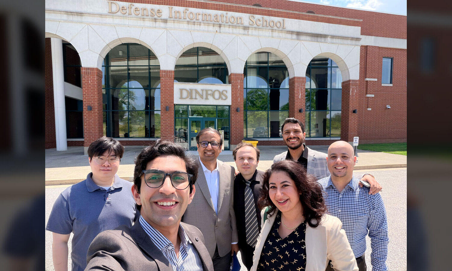 A photo of seven people taking a photo in front of a building. The building has letters that spell out \"Defense Information School.\"