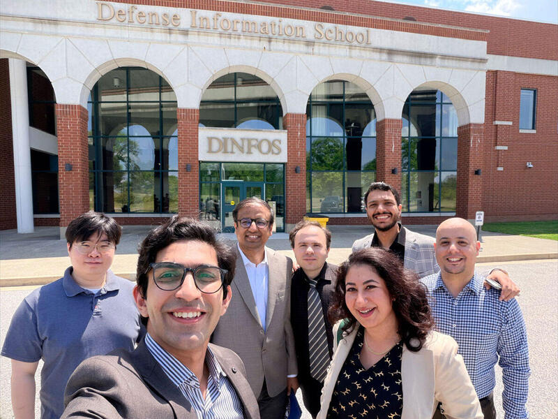A photo of seven people taking a photo in front of a building. The building has letters that spell out \"Defense Information School.\"
