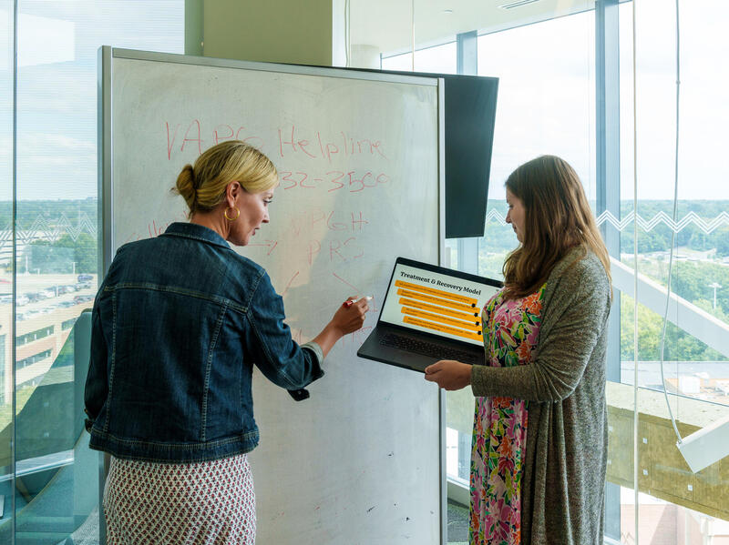 A photo of two women standing in front of a whiteboard. The woman on the left is holding a marker and writing on the board. The woman on the right is holding a laptop that both of the women are looking at. 