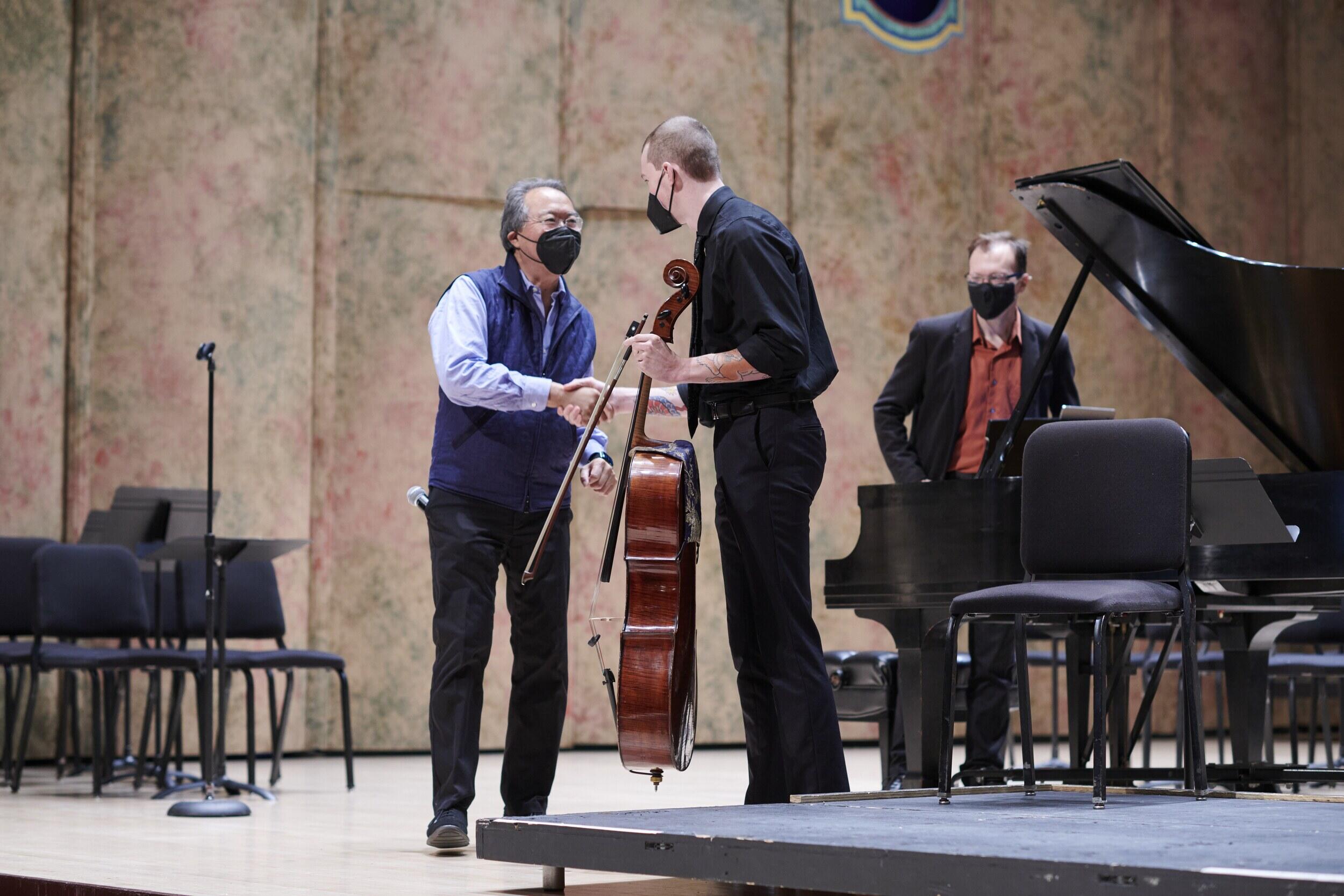 Two men searing face masks shaking hands in front of a piano. The man on the right is holding a cello. Behind them another man stands behind the piano. 