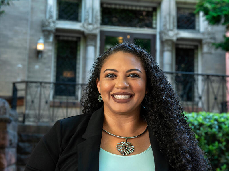 A woman in a green top and jacket smiles outside.