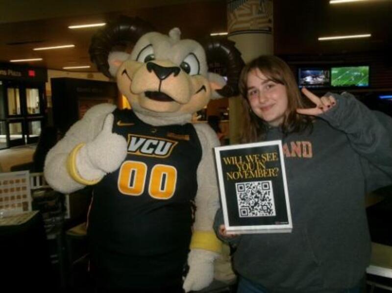 A photo of a woman standing to the right of Rodney the Ram. Rodney is giving a thumbs up and the woman is making a peace sign with her right hand. In her left hand she is holding a sheet of paper with a QR code and yellow text that reads \"WILL WE SEE YOU IN NOVEMBER?\" 