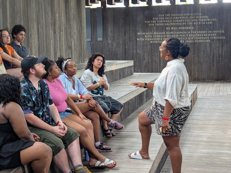 A photo of a woman talking to a crowd of seven people sitting on some steps. 