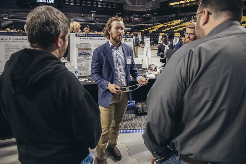 A photo of a man standing in front of a table with a poster board and holding a spliter. The man is talking to three other men who are standing around him in a semi-circle. 