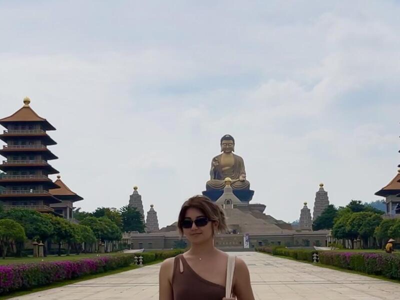 Naomi Ghahrai standing in the foreground on a pathway that leads to a large Buddhist monastery.