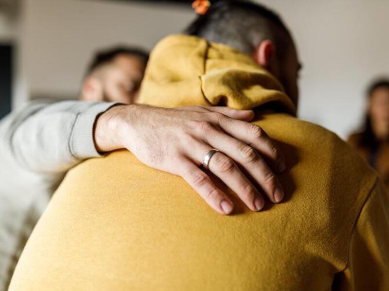 A photo of a man wearing a light gray sweat shirt sitting and comforting a man wearing a yellow hoodie next to him. The man in the gray sweashirt has his hand on the back of the man in the yellow hoodie. A burry woman sits across from them.