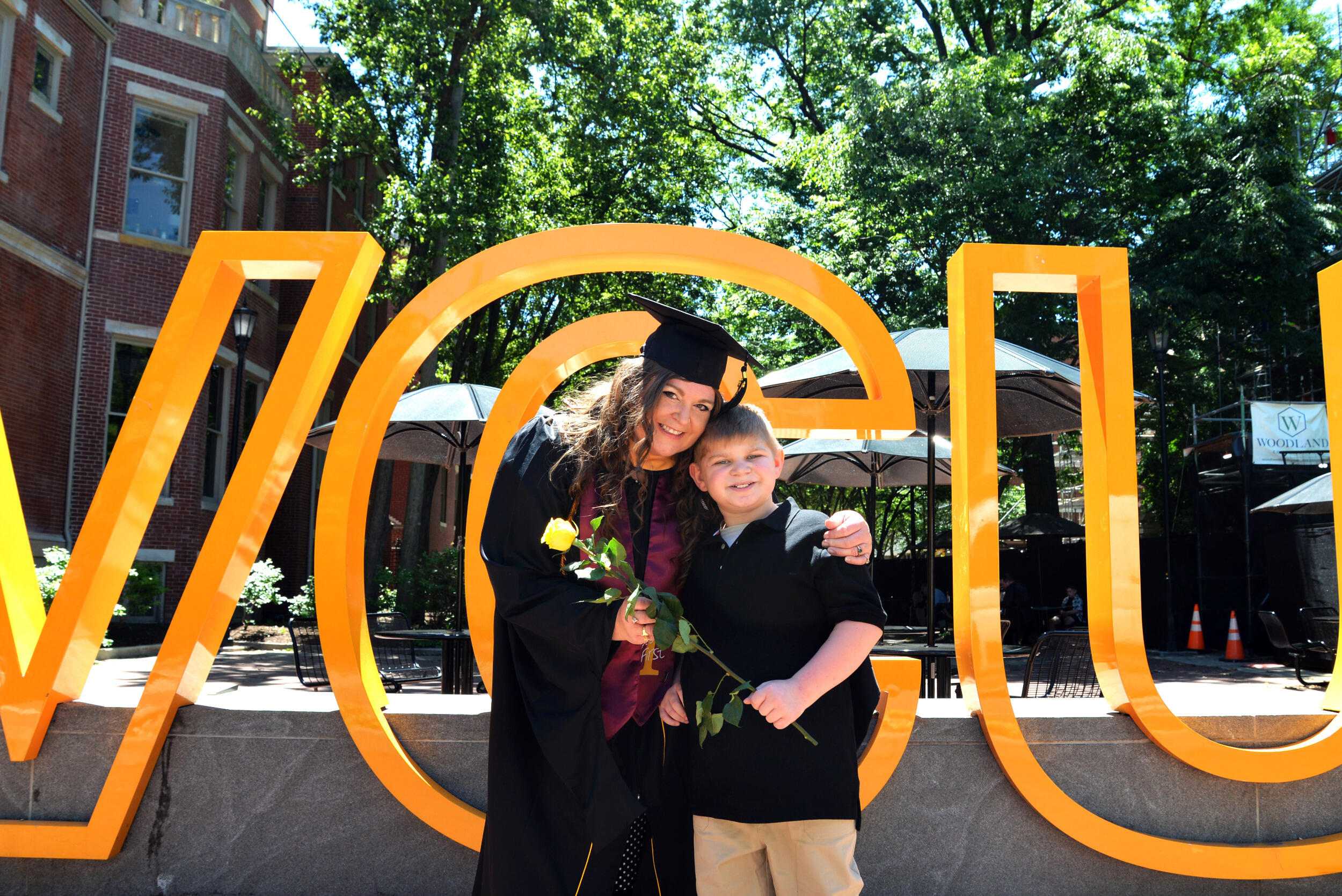 A smiling woman in a cap and gown has an arm over a smiling boy, and they hold a flower together.